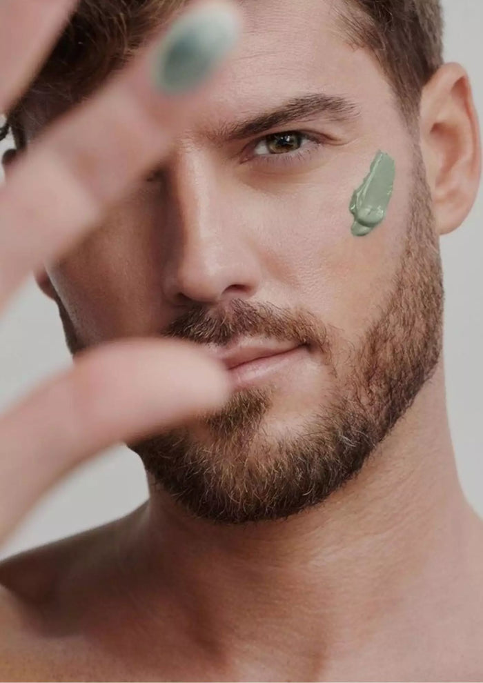 Homme barbu au regard intense appliquant une crème verte sur sa joue, main au premier plan, illustrant une routine de soin minimaliste et moderne pour hommes.